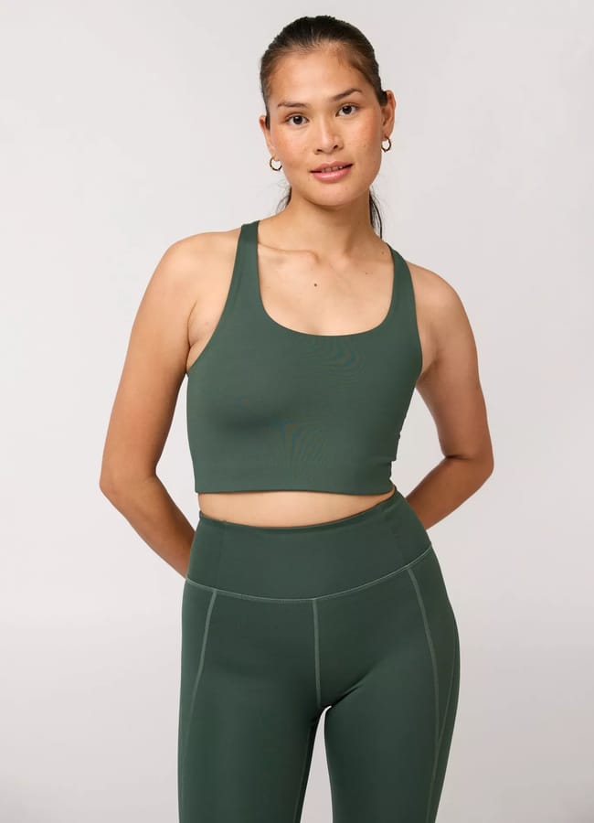 A person wearing a matching dark green athleisure sports bra and high-waisted leggings stands against a plain light background, arms behind their back and a slight smile.
