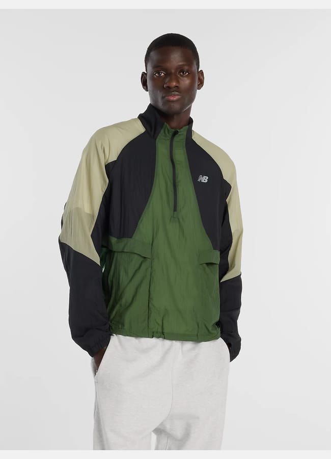A man stands against a plain white background, wearing gym clothes for men—a colour-blocked jacket in dark green, black, and light beige with a New Balance logo—and light grey sweatpants. His hands are in his pockets.