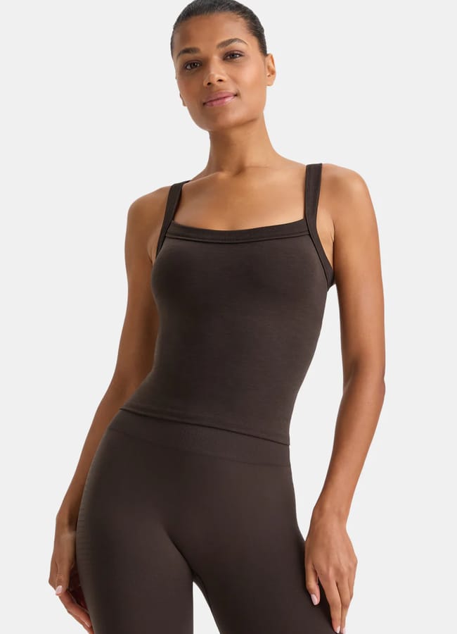 A woman stands facing forward in a fitted, dark brown athleisure vest top and matching high-waisted leggings. She has short hair and a neutral expression against a plain white background.