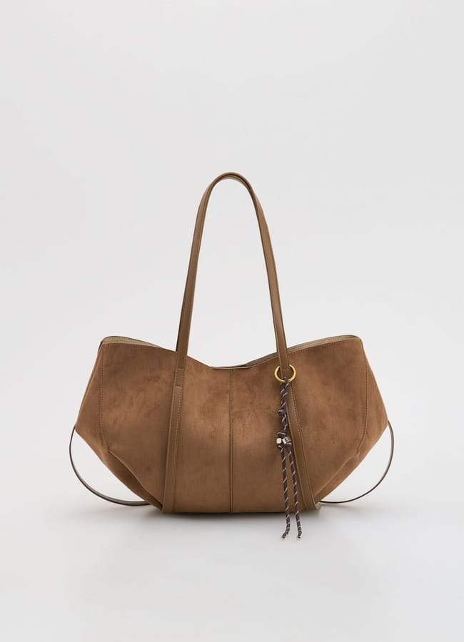 A brown suede tote bag with two long handles, subtle stitching, and a decorative tassel attached to a gold ring—an elegant essential for your no spend challenge, set against a plain white background.