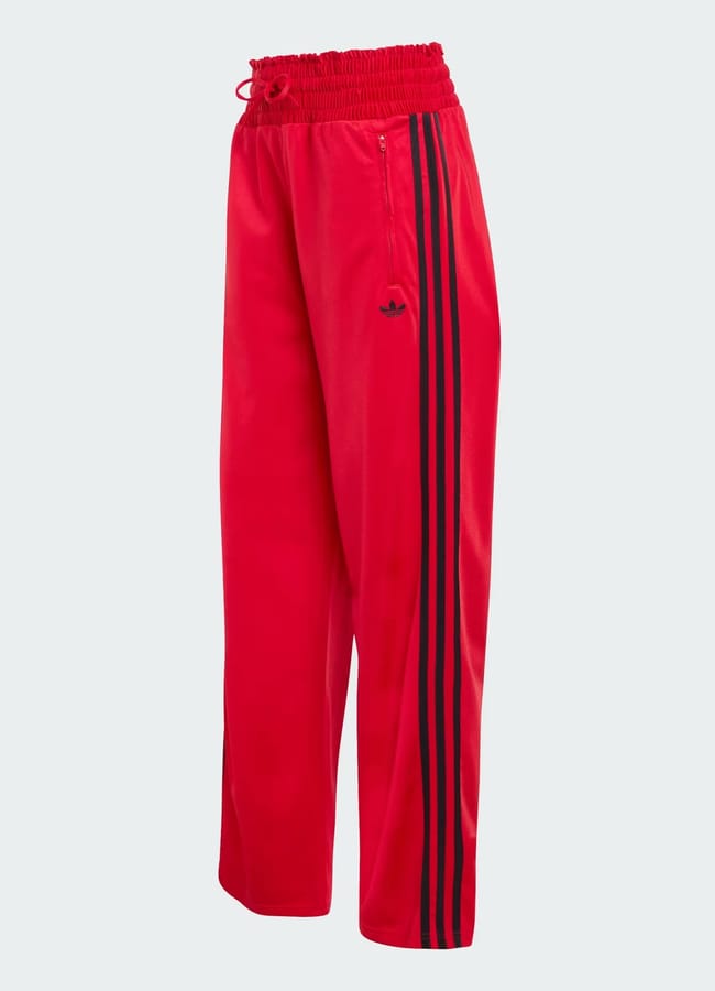 Bright red track trousers with an elastic waistband, drawstring, and black side stripes running down the legs. An embroidered black logo appears on the upper left thigh. These track trousers offer a relaxed, straight-leg fit.