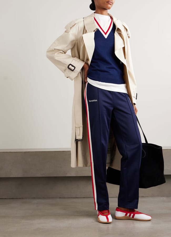 A person stands indoors wearing a beige trench coat over a navy V-neck jumper, white shirt, and blue athleisure track trousers with red and white stripes, paired with white trainers featuring red accents. They are holding a large black bag.
