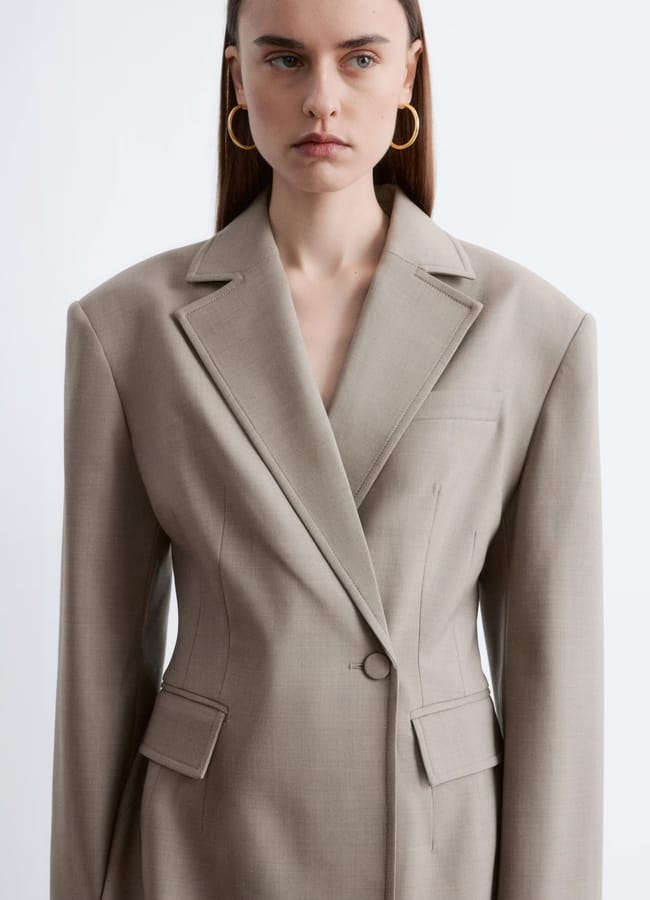 A woman wearing a structured, light beige blazer—one of the best blazers favoured by any fashion editor—stands against a plain white background. She has straight brown hair and gold hoop earrings, exemplifying chic high street blazers style.
