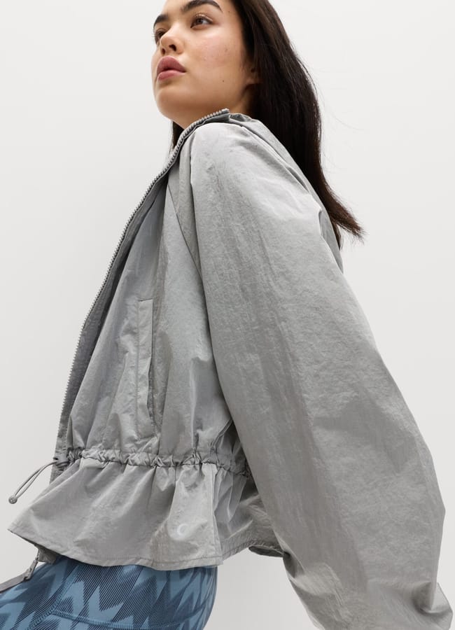 A woman with long dark hair wears a loose, light grey zip-up jacket with a cinched waist and patterned blue leggings—showcasing the latest activewear—while standing against a plain white background.