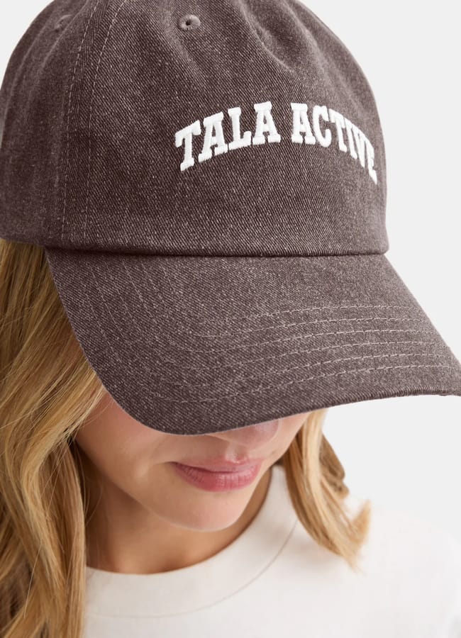A person wearing a dark grey athleisure baseball cap with the words TALA ACTIVE embroidered in white on the front. The person's face is partly obscured by the hat's brim, and they have fair, wavy hair.