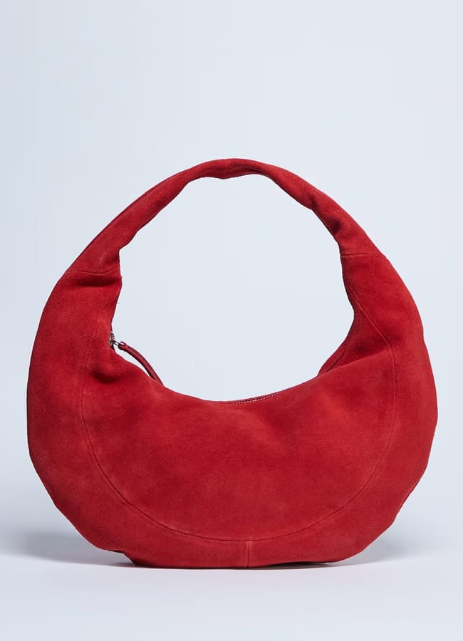 A round, red suede handbag with a single curved handle and a zip fastening, this chic piece is a standout among red accessories, displayed against a light grey background.