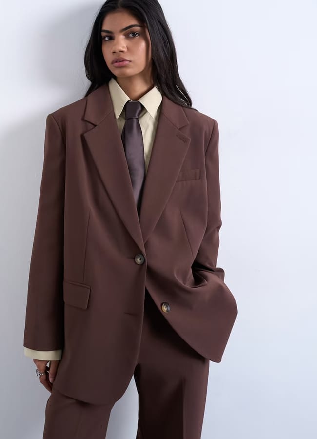 A person with long dark hair wears an oversized brown High Street Blazers suit with a matching tie and light beige shirt, standing against a plain, light-coloured wall—a look worthy of a Fashion Editor.