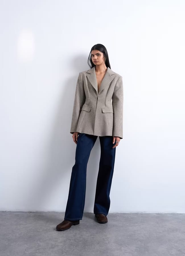 A woman stands against a plain white wall wearing a light grey High Street blazer, dark blue wide-leg jeans, and brown shoes. Her long dark hair is parted in the middle as she gazes slightly to the side, exuding fashion editor poise.