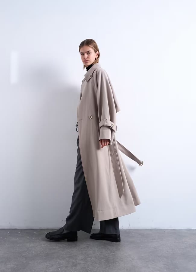 A person stands sideways against a plain white wall, wearing a long beige trench coat over dark trousers and black shoes—a stylish choice for casual outfits. The belt hangs loosely at the side, and the minimalistic space features a concrete floor.