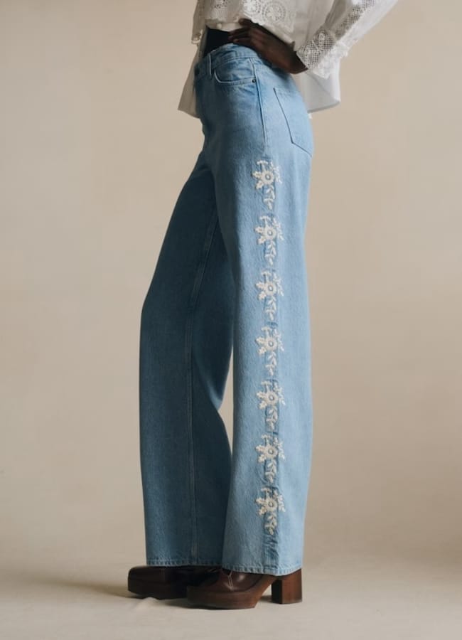 A person wearing high-waisted, light blue wide-leg jeans with white floral embroidery down the side seam, paired with a white lace blouse and brown platform shoes—an uplifting example of dopamine dressing—stands against a plain background.