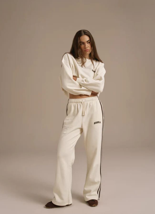 A woman stands with arms crossed, showcasing athleisure style in a loose-fitting, white long-sleeved sweatshirt and matching white tracksuit bottoms with black stripes down the sides, against a plain beige background.