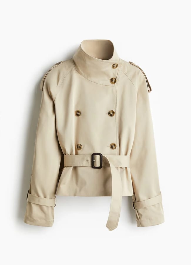 A beige cropped trench coat with a high collar, double-breasted button front, belted waist, and belted cuffs, styled effortlessly with track trousers and displayed against a plain white background.