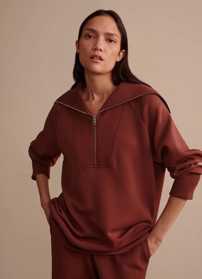 A person with long brown hair stands with hands in pockets, wearing a loose-fitting rust-coloured athleisure pullover with a front zip and matching trousers, against a plain beige background.
