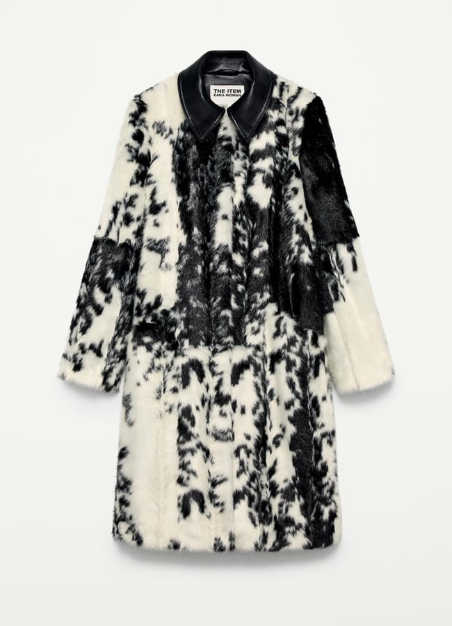 A long, faux fur coat with a black and white abstract, cow-like pattern channels dopamine dressing, featuring a black leather collar and button-up front, displayed against a plain white background.