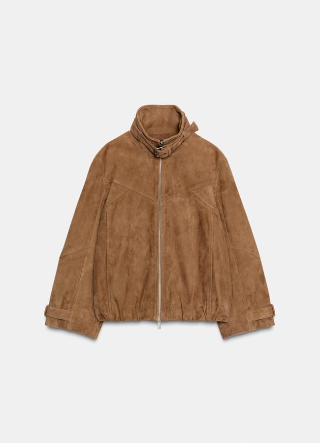 Light brown suede jacket with a high collar, front zip fastening, and adjustable strap at the collar. Perfect for pairing with track bottoms, it features long sleeves with strap details at the cuffs and a slightly loose fit. Displayed against a plain white background.