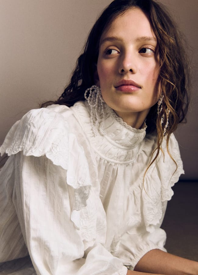 A young woman with wavy brown hair wears a high-neck, ruffled white blouse and large beaded earrings from the H&M new season collection. She looks thoughtfully to the side, posed against a neutral background.