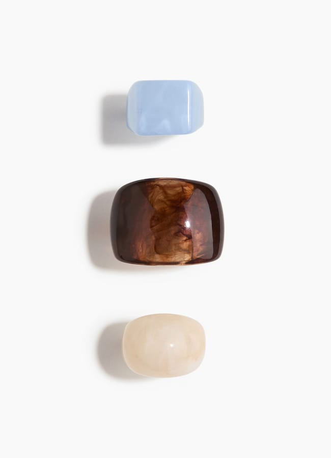 Three chunky rectangular rings from the H&M new season are arranged vertically on a white background. The top ring is light blue, the middle is brown and translucent, and the bottom one is off-white with a marbled appearance.