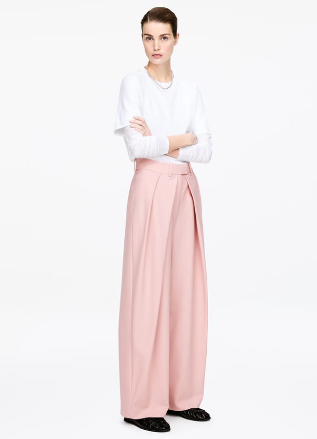 A person stands with arms crossed, wearing a white long-sleeved shirt, high-waisted pink pleated trousers with a matching belt, and black shoes against a plain white background.