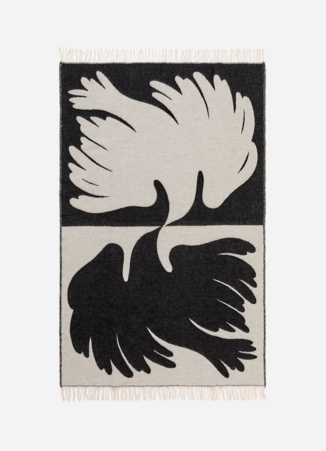 A high street homeware favourite, this woven blanket features two abstract bird-like shapes in a mirrored, yin-yang style—white on black above and black on white below—with short fringe on both edges.