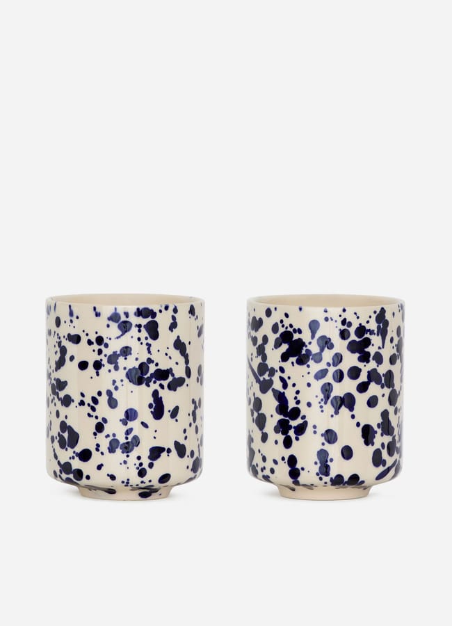 Two round ceramic cups with a cream base colour, decorated with irregular, dark blue splatter patterns, offer a stylish touch to your high street homeware collection. They’re placed side by side on a plain white background.