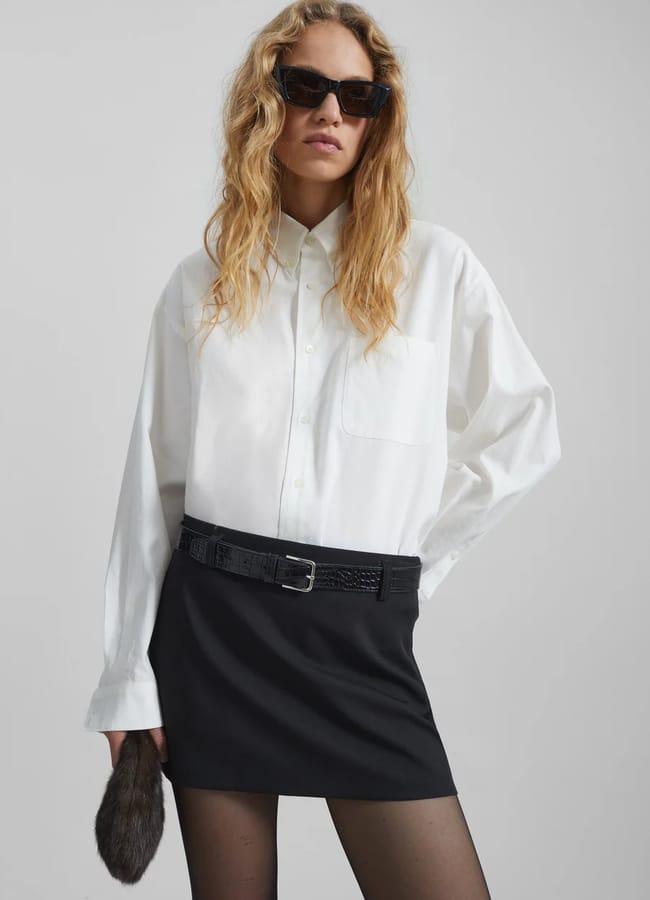 A woman with wavy blonde hair wears black sunglasses, a loose white shirt, black mini skirt, and tights. Holding a furry accessory, she stands confidently against a plain light grey background. Perfect styling inspiration for fans of white shirts.