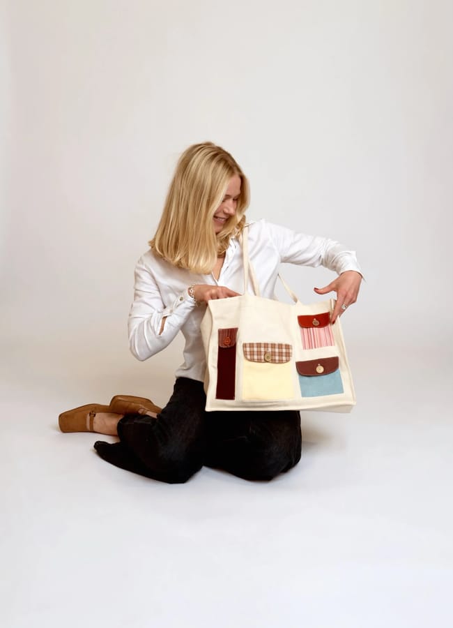 A woman with blonde hair, wearing a white shirt and black trousers, sits on the floor smiling whilst holding up a large tote bag with colourful patchwork pockets—perfect for your February pay day treat—against a plain white background.
