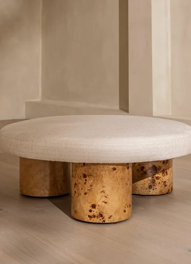A modern, low-profile ottoman with a round, beige upholstered cushion and three cylindrical wooden legs, set against a minimalist, light-coloured wall and floor.