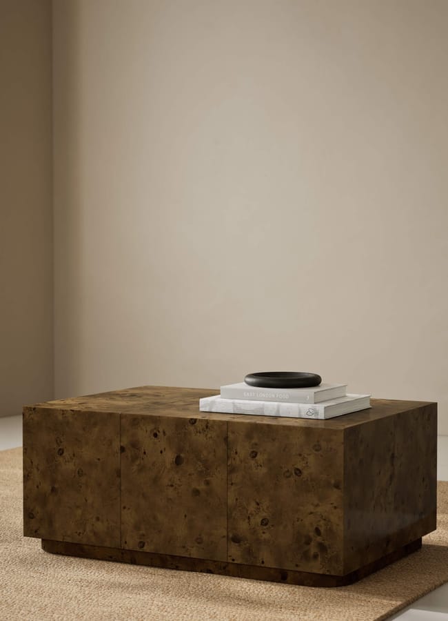 A square wooden coffee table with a rich, brown finish sits on a tan rug. On top of the table are two stacked books and a small black dish, set against a neutral beige wall.