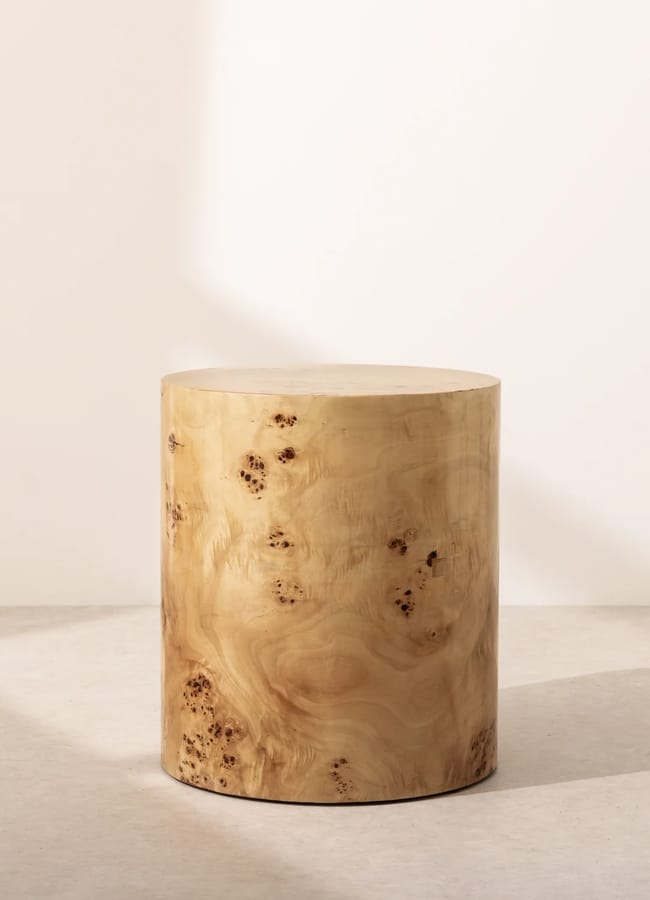 A cylindrical wooden stool with a smooth, light-brown finish and natural wood grain patterns sits on a light-coloured floor against a plain, off-white wall.