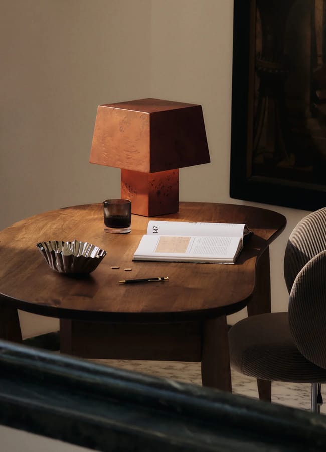 A wooden table with an open book, a pen, a glass, a decorative bowl, and a geometric brown lamp sits beside a curved, upholstered chair in a softly lit room.