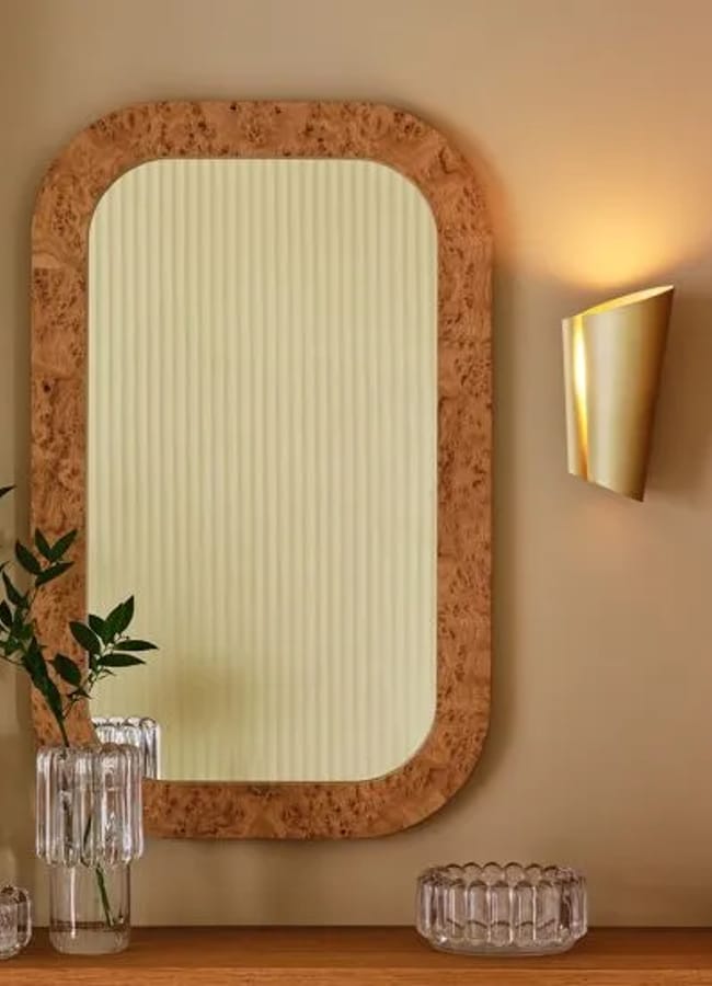 A rectangular mirror with a rounded wooden frame hangs on a beige wall, next to a lit gold wall sconce. A glass vase with green branches and two glass dishes are placed on a wooden shelf below.