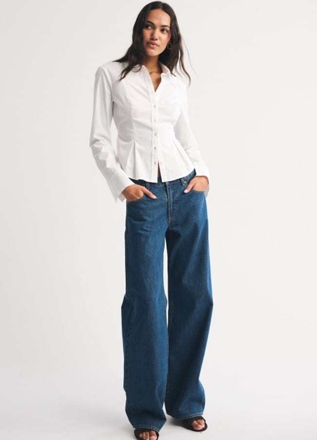 A woman stands against a white background wearing a fitted white shirt, wide-leg blue jeans, and black shoes, her hands in her pockets and a relaxed expression. This look highlights the timeless appeal of classic white shirts.