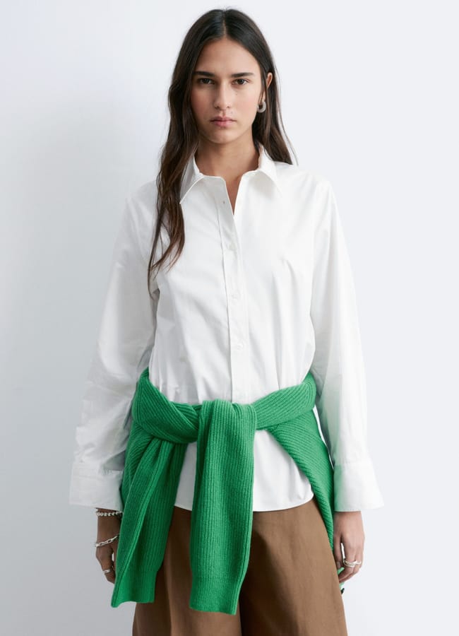 A woman with long dark hair wears white shirts and brown trousers, with a green jumper tied around her waist. She stands against a plain white background, looking directly at the camera.