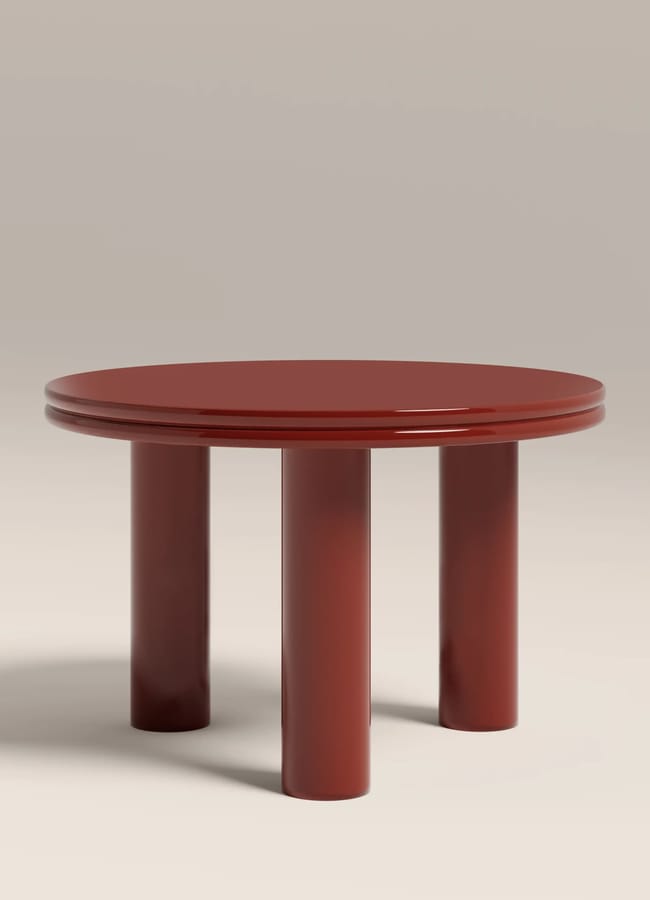 A round, glossy, dark red table with three thick cylindrical legs adds a bold touch to red and pink interiors, set against a plain beige background.