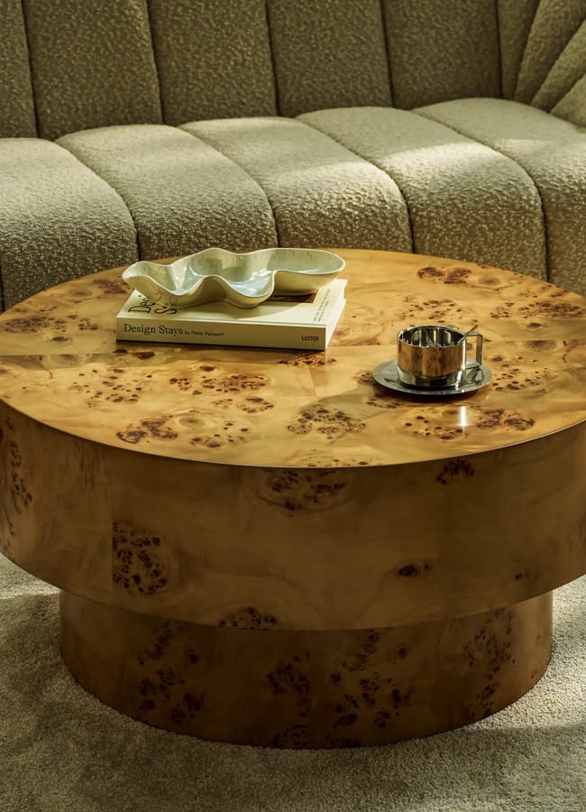 A round wooden coffee table with a glossy, burr wood finish holds a stack of books, a ceramic decorative dish, and a metallic cup. The table sits on a light carpet in front of a textured beige sofa.