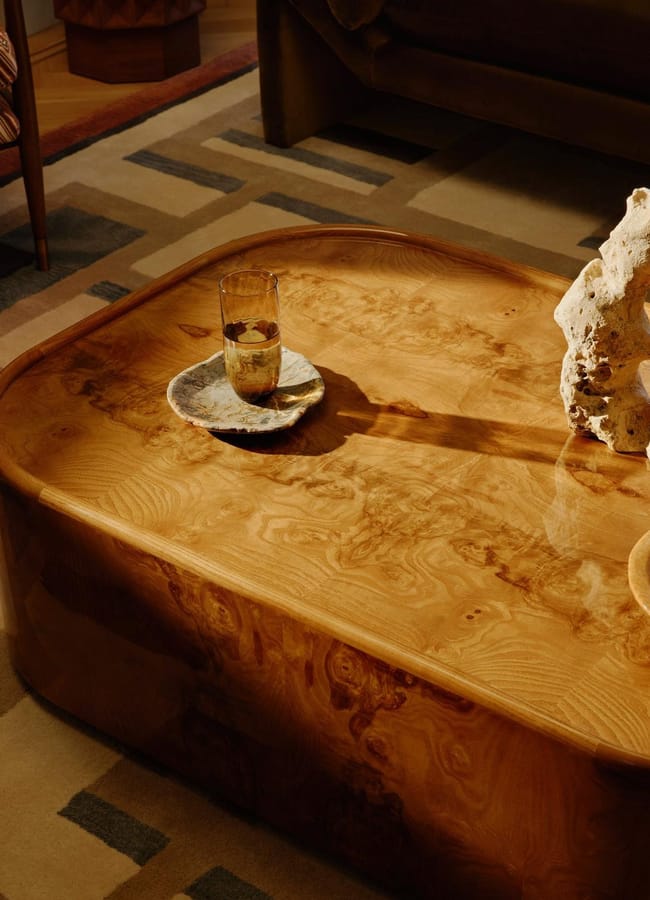 A glossy wooden coffee table holds a glass of drink on a saucer, a decorative coral-like sculpture, and a wooden tray with small objects. The table sits on a patterned rug in a cosy living room.