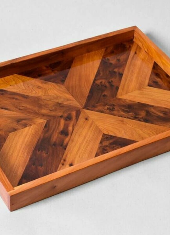 A rectangular wooden tray with raised edges featuring a geometric, starburst pattern made from various wood tones and grains. The tray is photographed on a light grey surface.