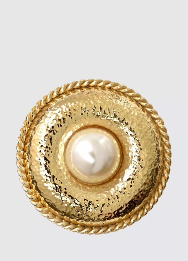 A round, gold brooch with a hammered texture and central white pearl-like stone. The twisted rope border adds timeless style, making it a refined fashion accessory against a plain light grey background.