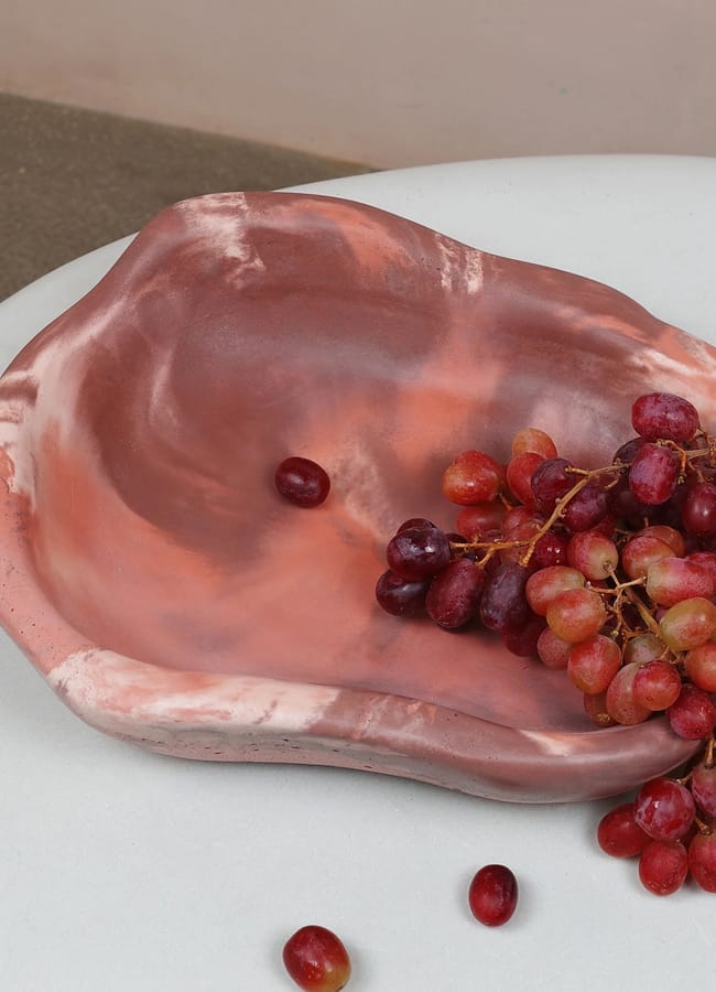 A marbled pink and white ceramic bowl with an irregular shape, perfect for red and pink interiors, holds a bunch of red grapes on a smooth white surface. One loose grape sits inside the bowl and another rests nearby.
