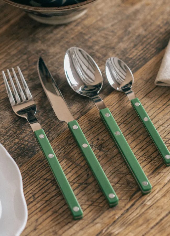 A fork, knife, tablespoon, and teaspoon with green handles from high street homeware are neatly arranged on a rustic wooden table next to beige serviettes and a white plate.