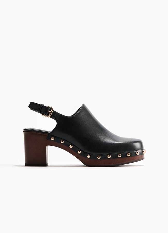 A black leather clog from the H&M new season collection, featuring a thick brown heel, gold studs along the sole, and an adjustable ankle strap with a gold buckle, shown on a white background.
