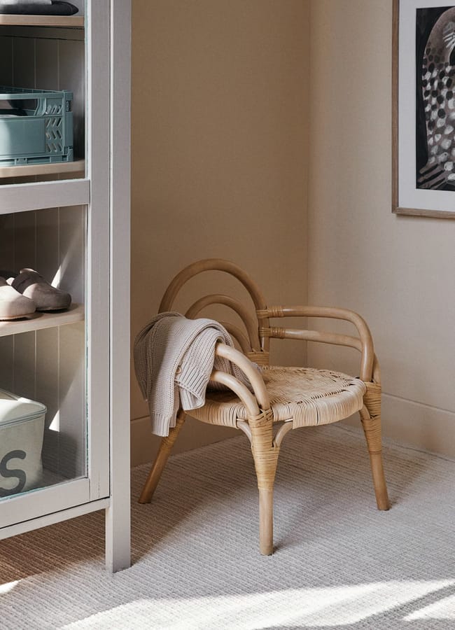 A woven rattan chair with curved armrests sits on a light carpeted floor, draped with a folded beige jumper. Nearby, a shelving unit holds high street homeware storage baskets, and a framed feather artwork hangs on the wall.