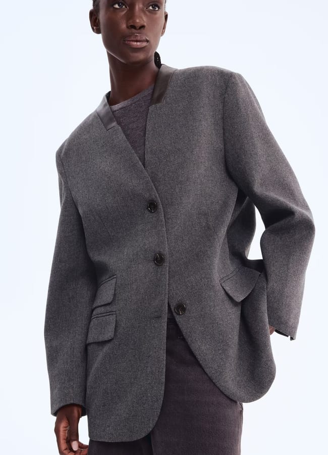A person wearing a loose-fitting, grey blazer with black buttons and a black collar detail over a dark shirt and trousers, channelling a Wuthering Heights wardrobe vibe, poses with one hand in a pocket against a plain light background.
