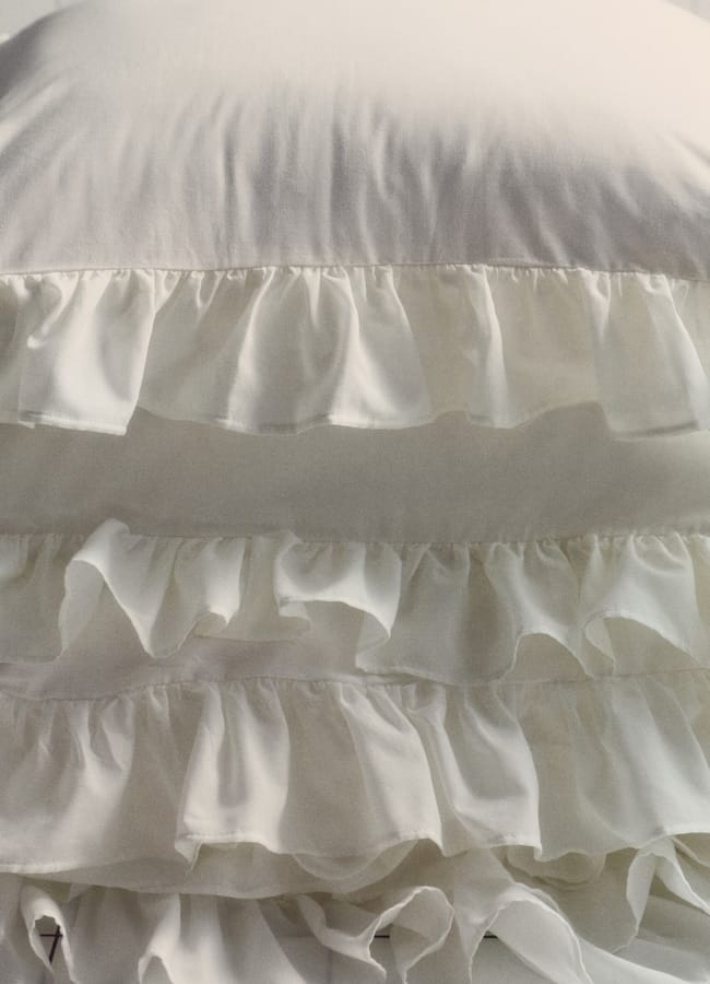 A close-up of a white pillow with several layers of ruffled fabric, sitting on a white tiled surface. The soft, decorative texture brings a touch of high street homeware style to any space.