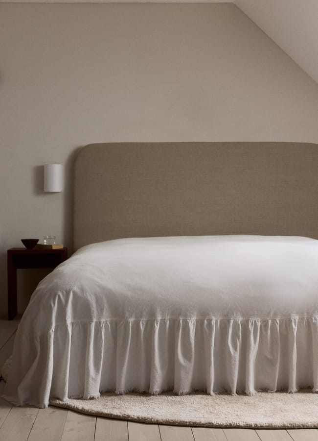 A minimal, cosy bedroom with a large beige upholstered headboard, white bedding with a ruffled skirt, and touches of high street homeware on a small wooden bedside table—all set against light wood flooring.