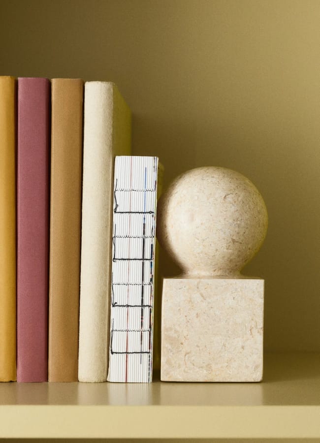 A row of books with neutral-coloured covers stands on a beige shelf next to a decorative stone sphere, creating a stylish scene reminiscent of high street homeware displays. The background is a matching beige colour.