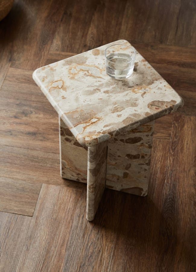 A small, square, stone side table with a speckled beige and brown surface—perfect for adding high street homeware style—sits on a wooden floor. A glass of water rests on top of the table.