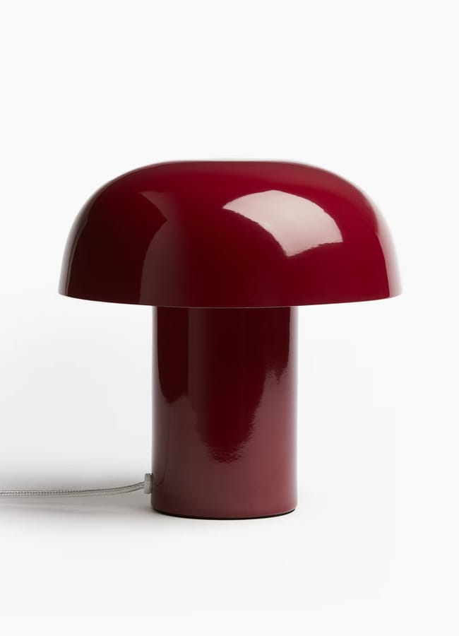 A modern table lamp with a glossy, dark red mushroom-shaped design complements red and pink interiors, featuring a rounded dome top and cylindrical base, set against a plain white background. A power lead extends from the base.