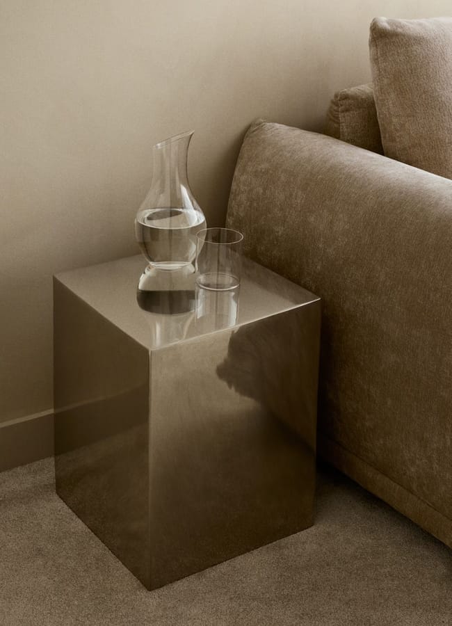 A metallic cube side table from high street homeware holds a clear glass carafe filled with water and an empty glass tumbler, placed next to a beige upholstered sofa on a carpeted floor in a minimalist room.