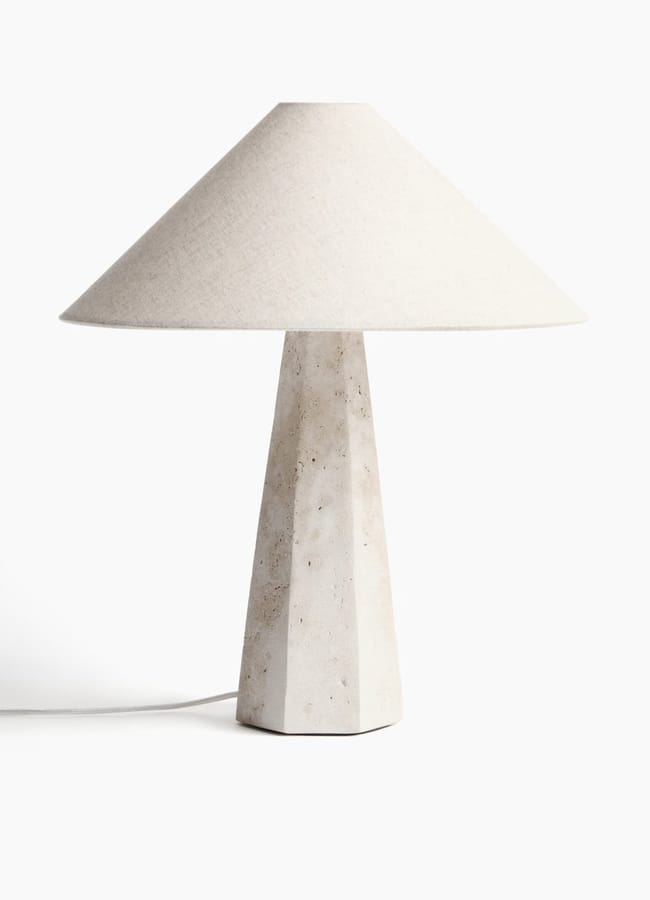 A modern table lamp with a geometric, stone-like base and a wide, conical off-white fabric lampshade, echoing high street homeware trends, set against a plain white background.